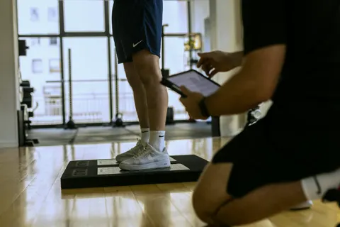 Stopy zawodnika na platformach pomiarowych, kadr od bioder w dół. Karol klęczy obok i ustawia test na tablecie.