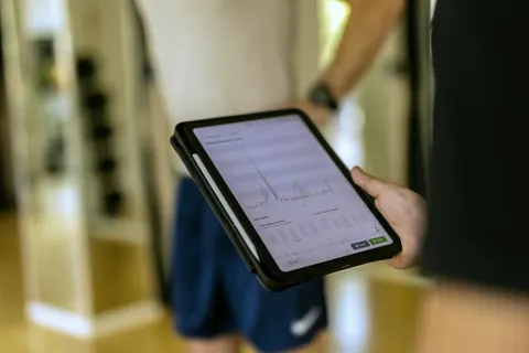 Zbliżenie na tablet z wynikami testu skoku trzymany przez Karola, w tle rozmyta sylwetka zawodnika.